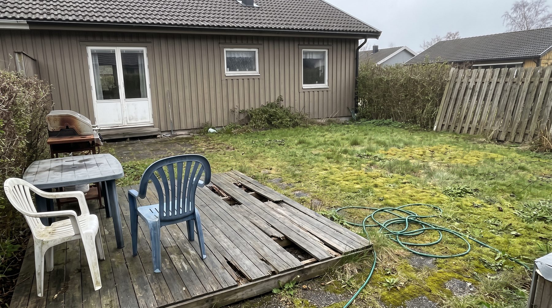 Rutten trätrall med plastmöbler och mossig gräsmatta bakom ett hus med brun panel.